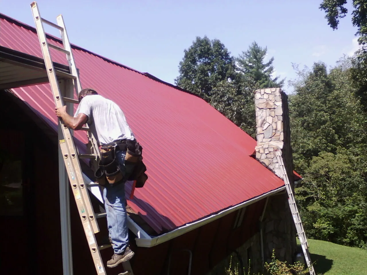 Roofer on a ladder beside a red metal roof and stone chimney — Soffit & Fascia Repair by Roofing Brothers in Swampscott